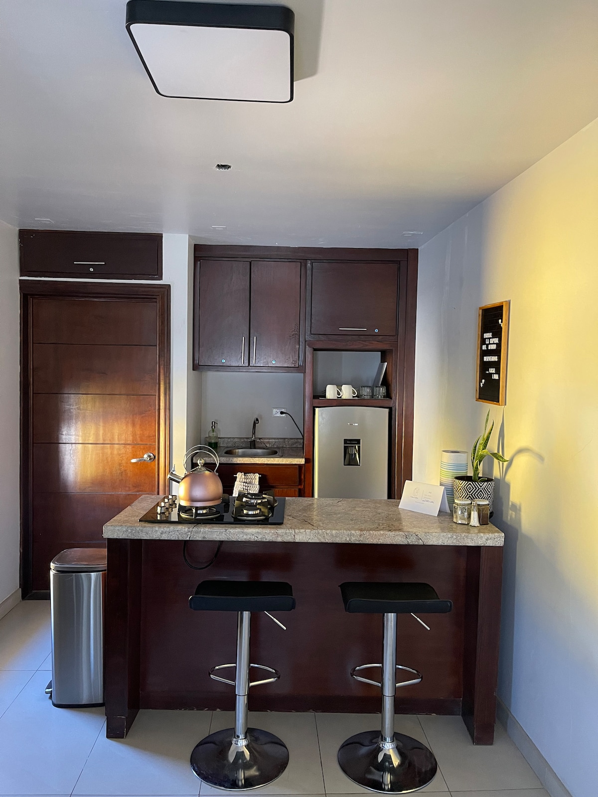 A compact kitchen area is featured, highlighted by a sleek granite countertop with two bar stools. Cabinetry provides ample storage, and stainless steel appliances, including a refrigerator and stove, are visible. Natural light enhances the warm tones of the space.