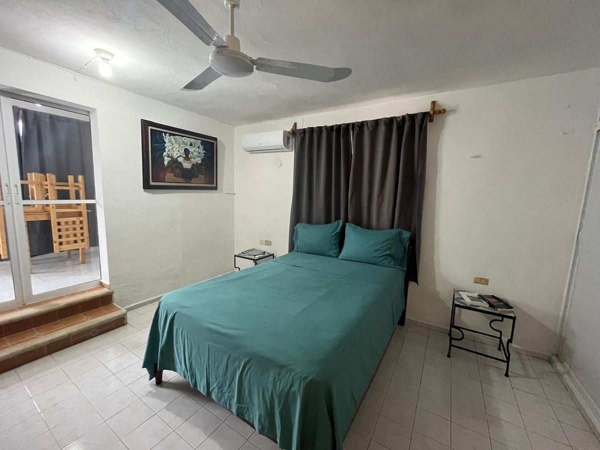 A well-organized room features a spacious bed with a teal coverlet and matching pillows. Natural light filters in from glass doors leading to an outdoor area. A small table is situated next to the bed, while dark curtains provide privacy.