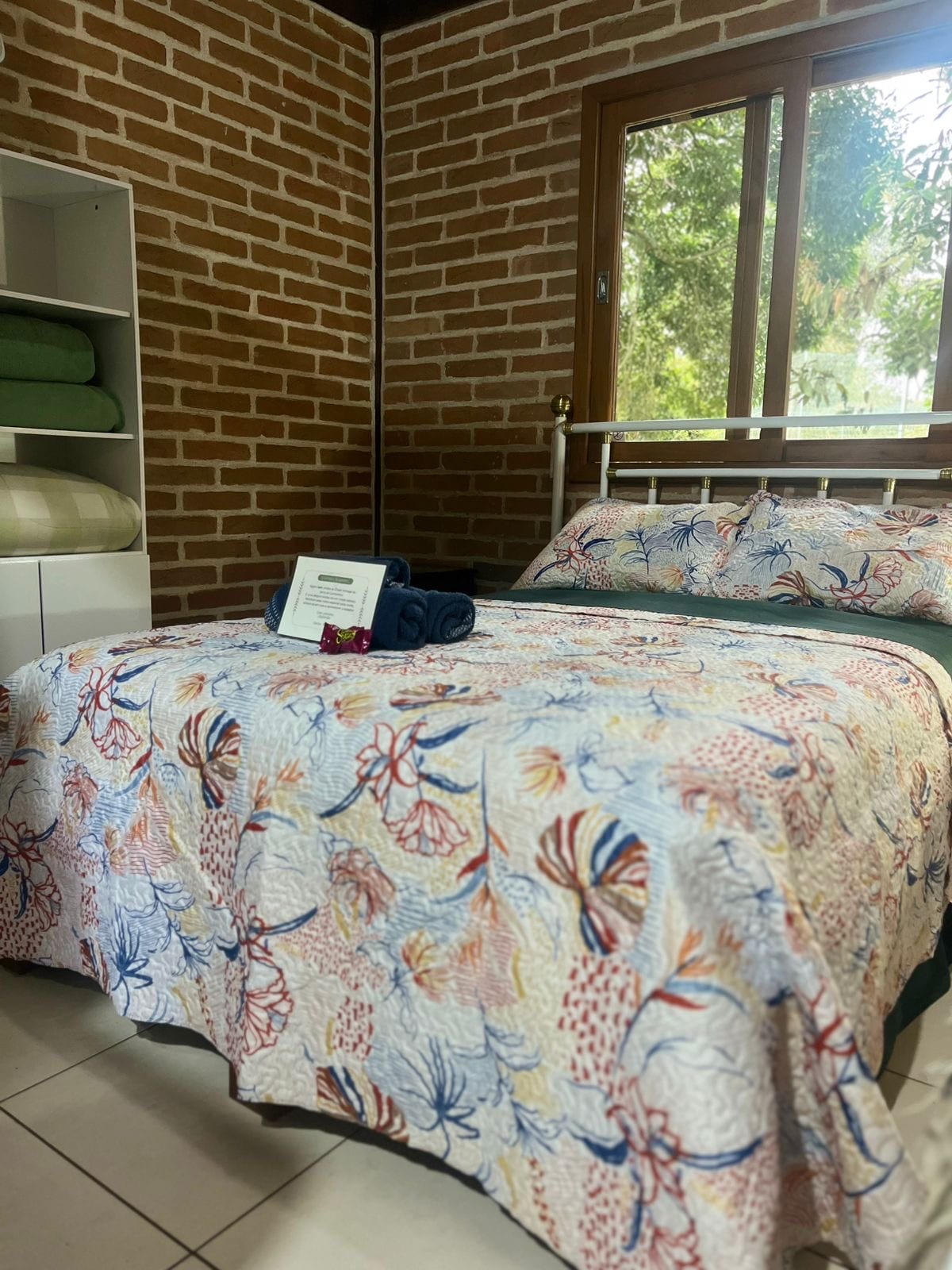 A cozy sleeping area features a bed covered with a floral bedspread, accompanied by neatly arranged towels. A brick wall adds texture, while natural light filters through a nearby window, offering a view of the surrounding greenery.