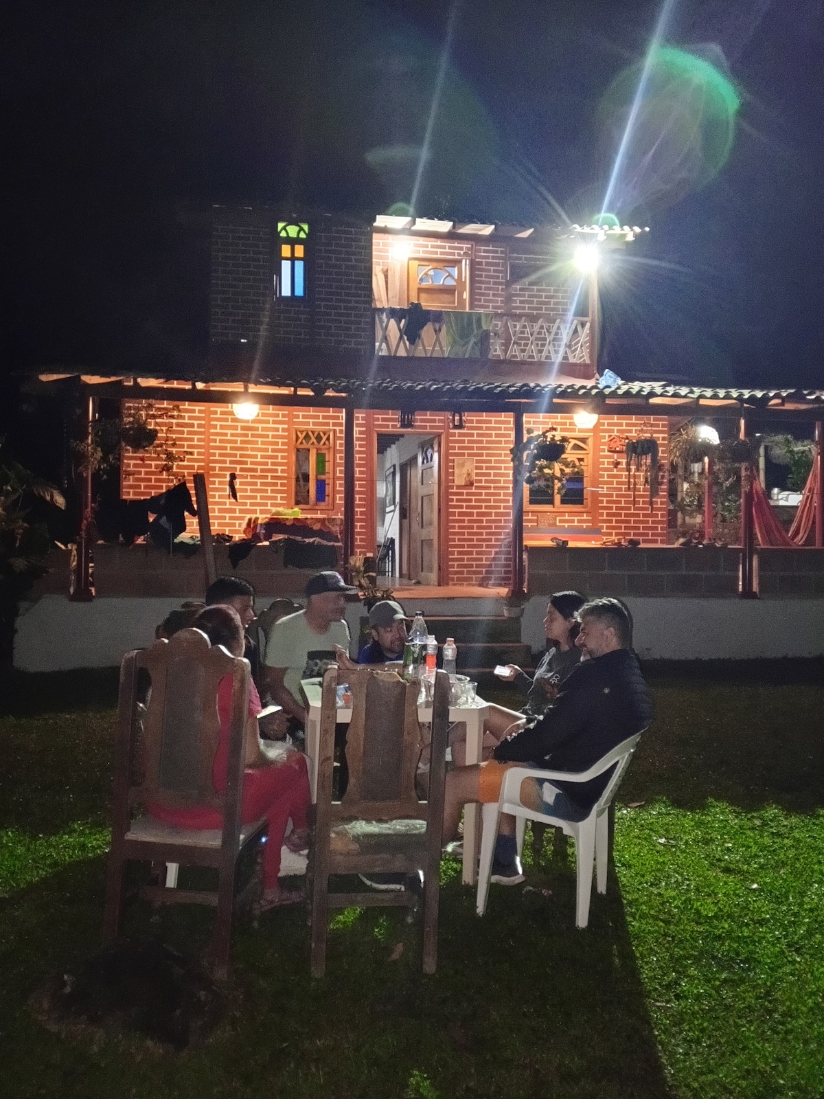 A cozy gathering is taking place outdoors, with a group of individuals seated around a large table. The warm glow of lights illuminates the rustic brick cabin in the background, which features a balcony and wooden railings. Lush green grass surrounds the scene, enhancing the inviting atmosphere.