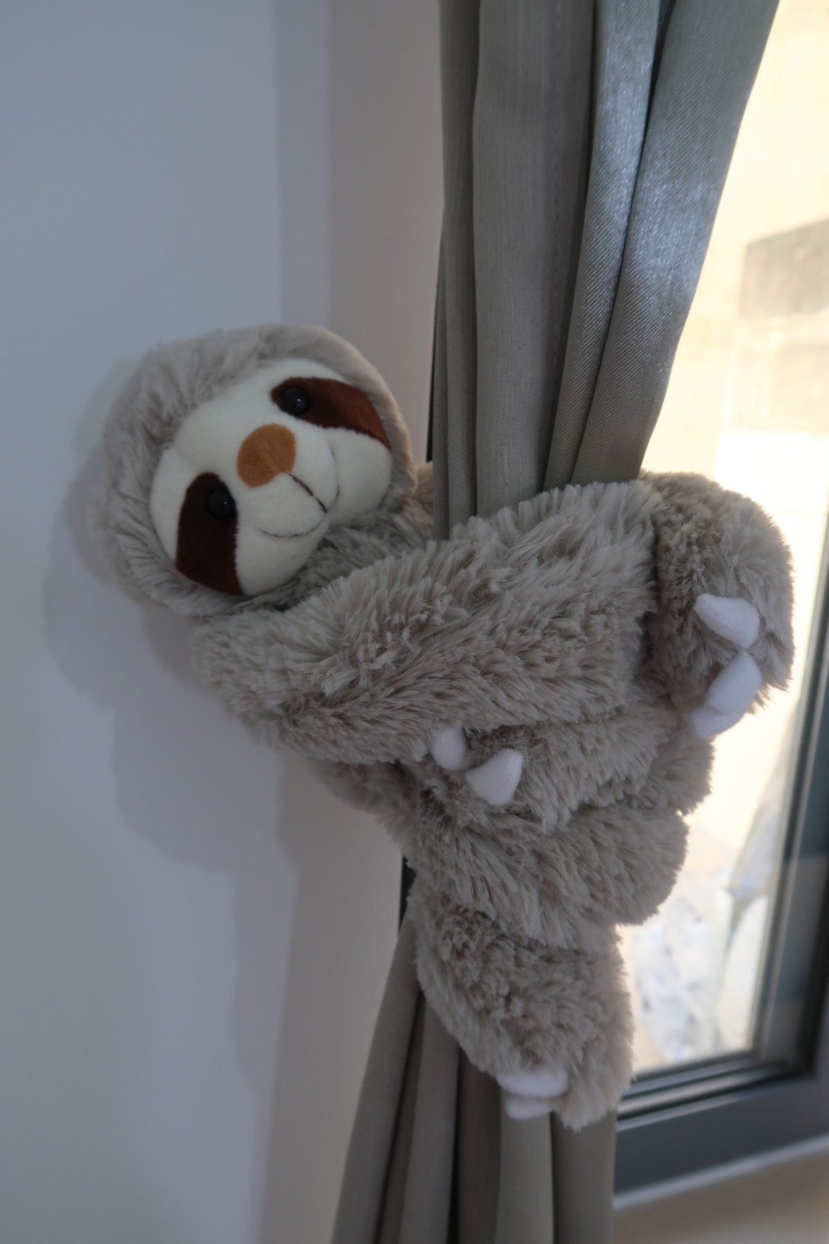 A soft, plush sloth toy is nestled among light curtains, with its head and arms playfully peeking out. The sloth's facial features are detailed, showcasing a gentle expression that adds a touch of whimsy to the room.