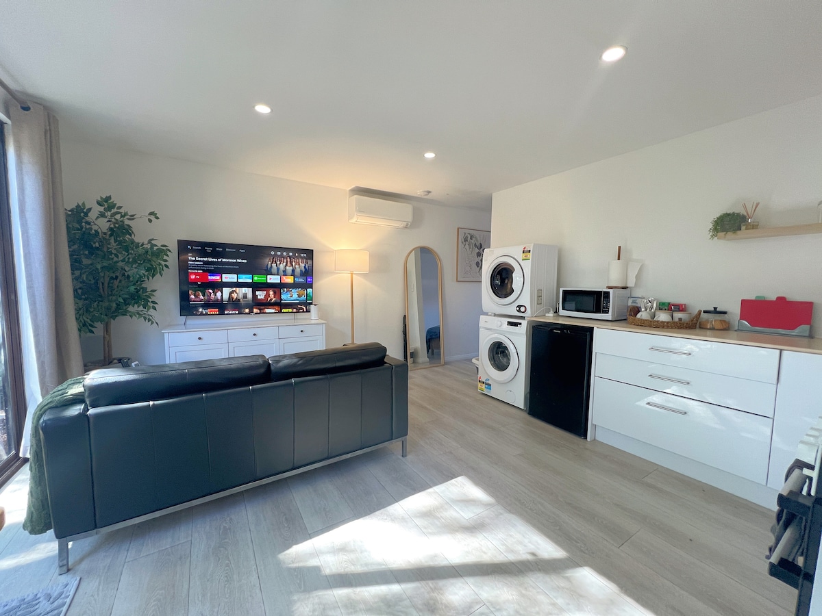 A bright living space features sleek white cabinetry and modern appliances, including a washer-dryer and microwave. A dark sectional sofa faces a large television, while natural light streams in from the windows. A small decorative table adds a touch of style to the room.