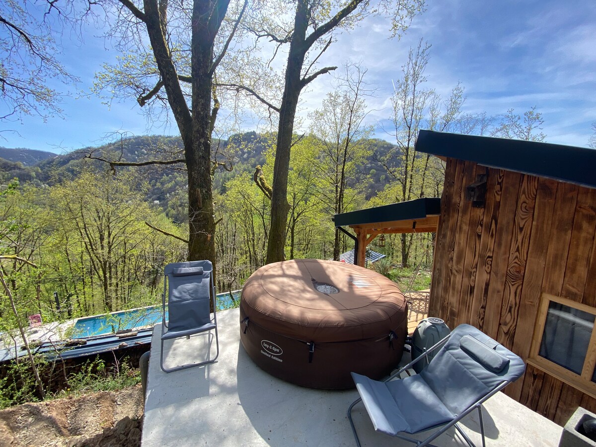 An outdoor space features a round jacuzzi surrounded by trees and greenery. Two lightweight chairs are positioned nearby, providing a place to relax. A scenic landscape of rolling hills under a clear blue sky adds a tranquil backdrop.