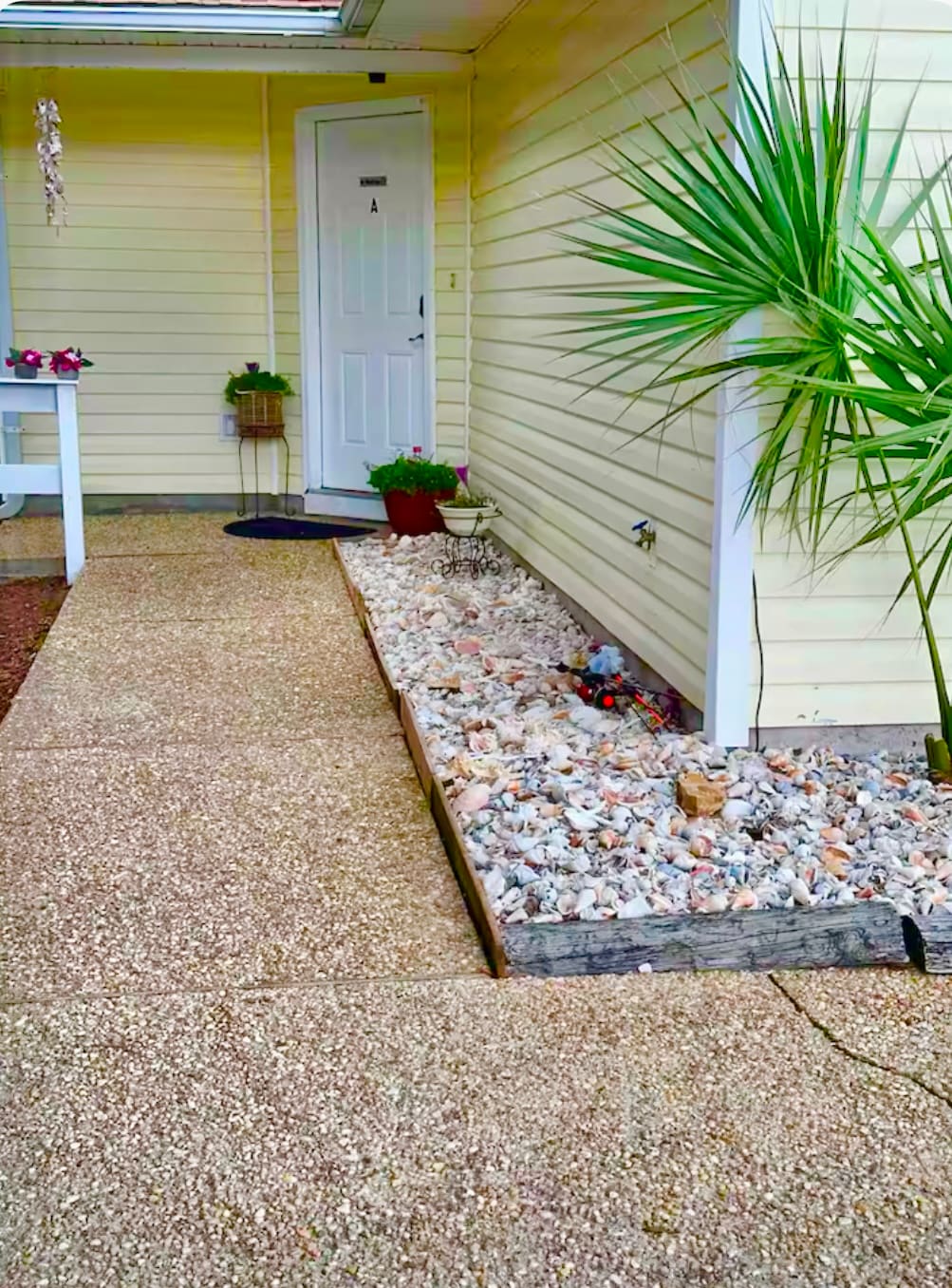A pathway lined with pebbles and seashells leads to a door framed by a small potted palm and colorful plants. The entrance features a light-colored exterior wall with a single door and a welcome mat, providing an inviting approach.
