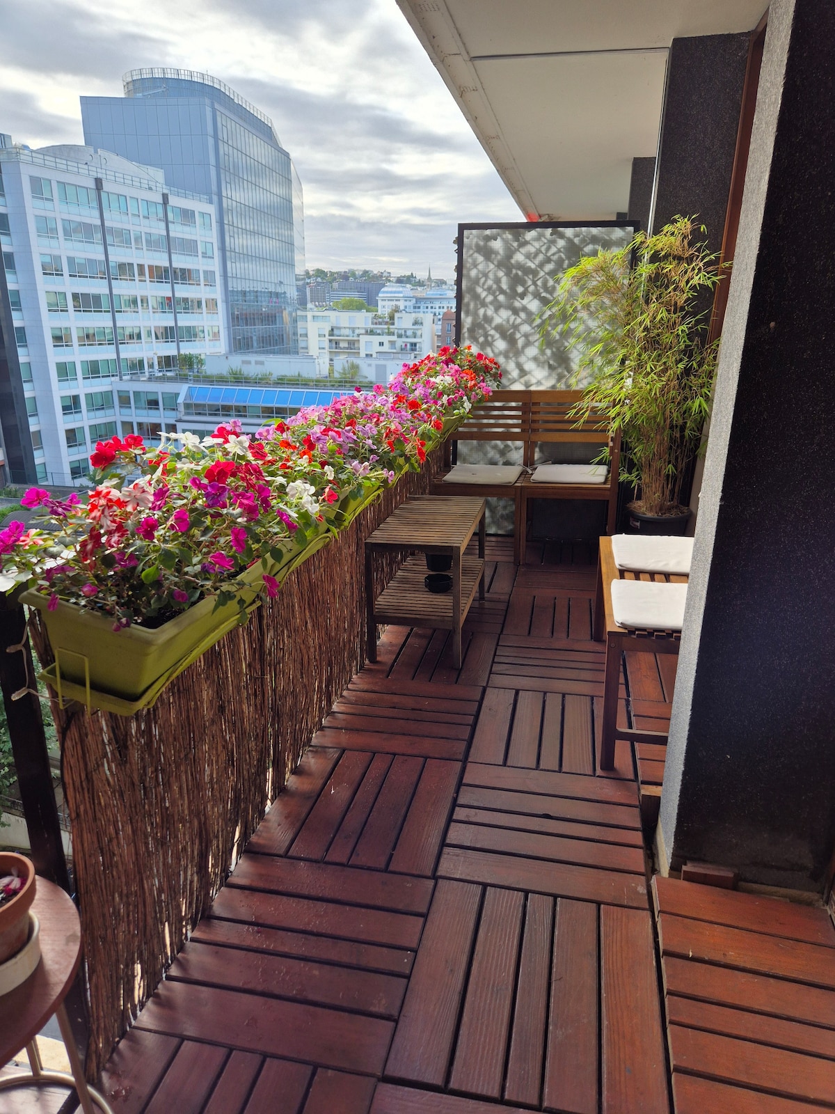 A private balcony is adorned with colorful flowers in various planters, creating a vibrant atmosphere. Wooden flooring extends throughout the space, which includes a small table and chairs, providing a comfortable seating area to enjoy the view of the surrounding cityscape.