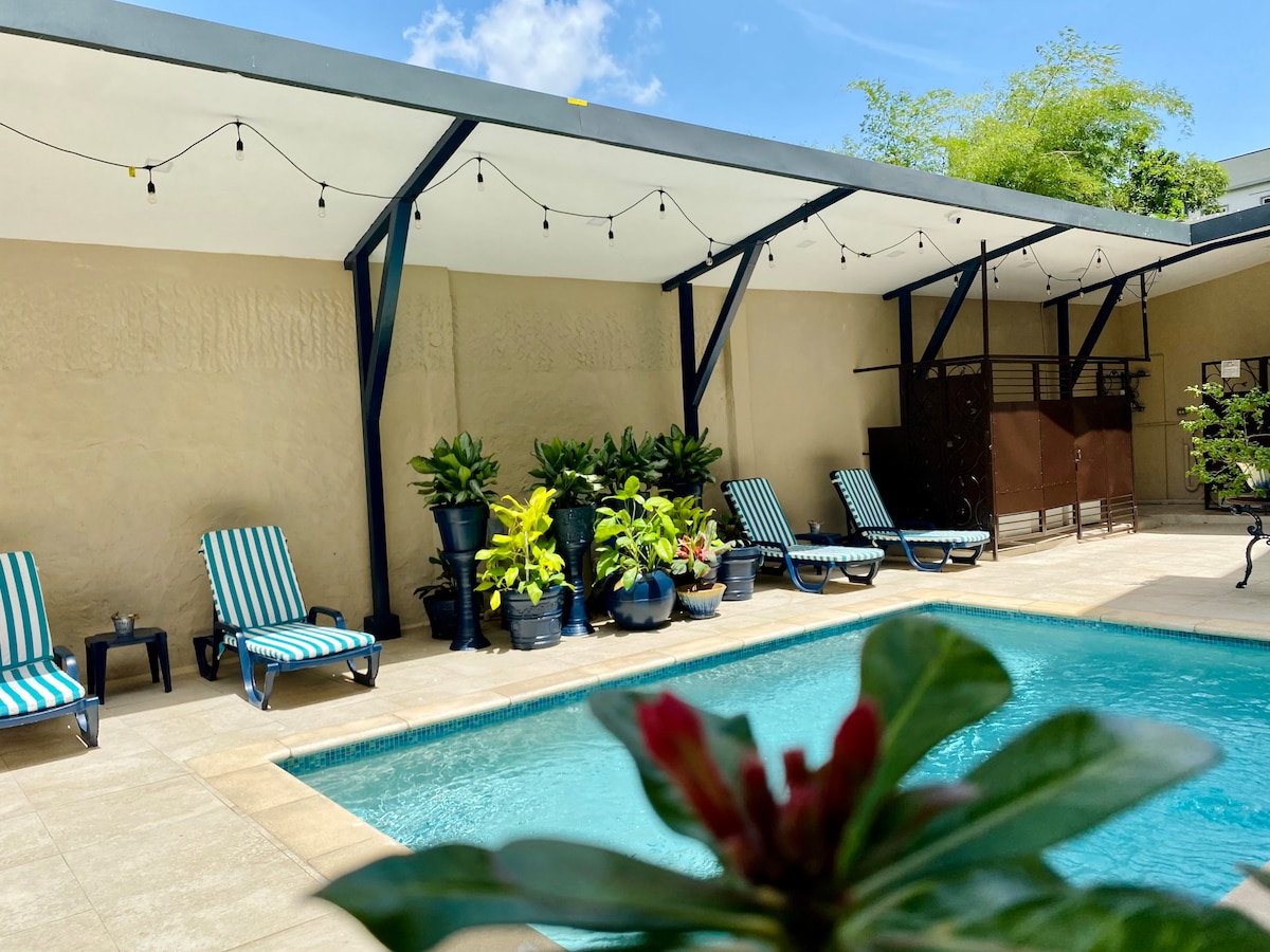 A pool area is highlighted by a clear blue water surface and surrounded by several lounge chairs with striped cushions. Decorative plants in black pots are positioned around the space. A canopy overhead offers shade, with string lights hanging above for a warm ambiance.