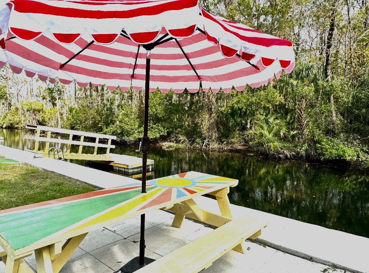 A vibrant picnic table with a sunburst design is positioned next to a calm canal. The area is shaded by a large red-and-white striped umbrella, providing a comfortable spot for relaxation. Lush greenery lines the banks, enhancing the natural setting.