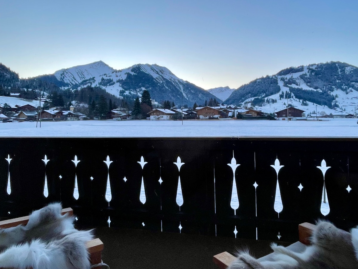 A scenic view from the balcony captures the snow-covered landscape and the surrounding mountains. Traditional wooden chalets dot the valley below, framed by the silhouette of distant peaks under a soft blue twilight sky.
