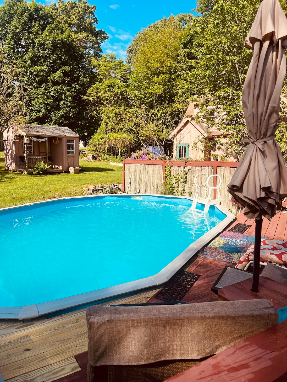 A serene outdoor space features a clear blue pool surrounded by wooden decking and soft seating. Lush green trees frame the area, with a charming garden shed and colorful plants in the background, creating a peaceful and inviting atmosphere.