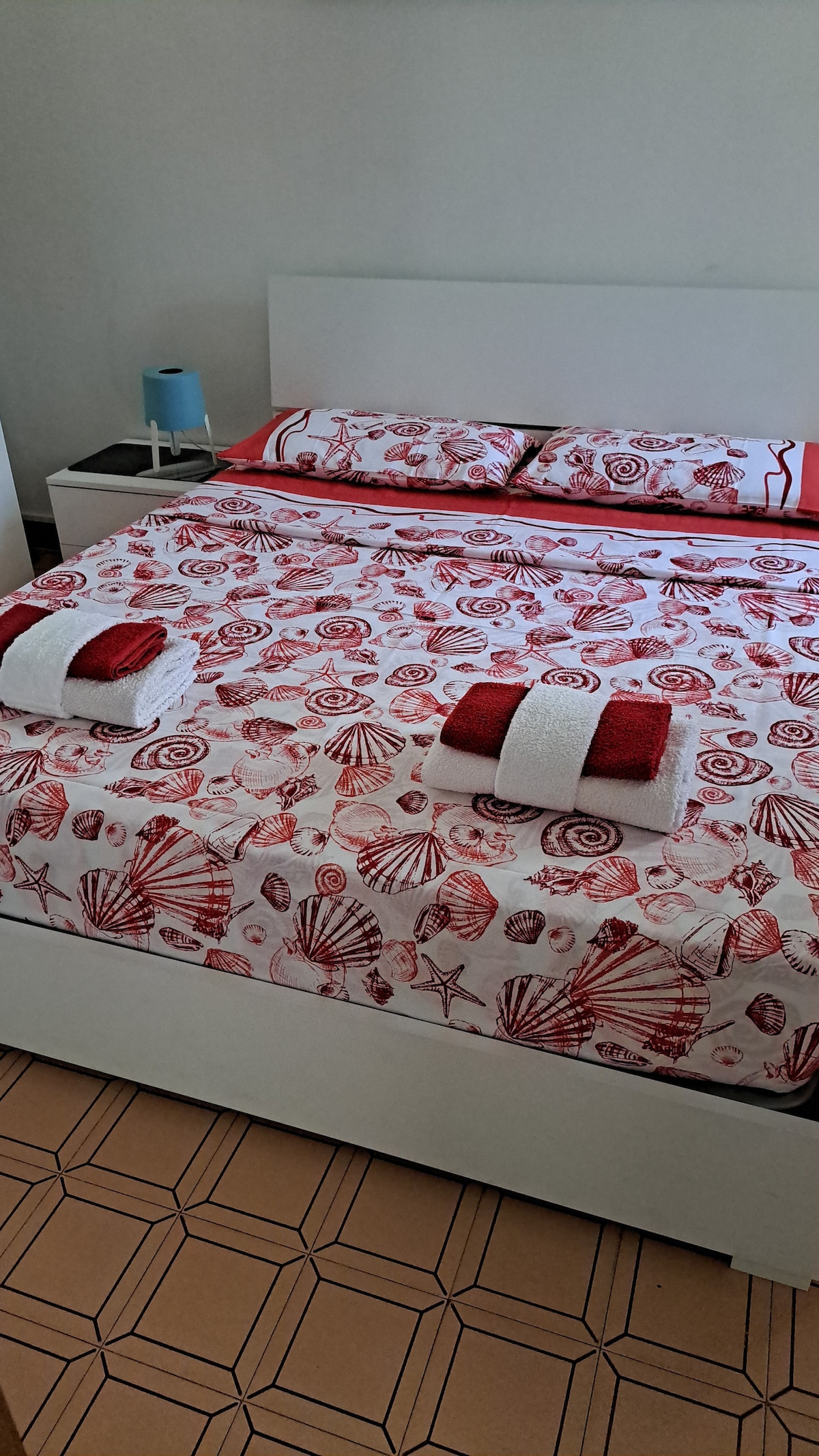A spacious bed is dressed in a bright red and white shell-patterned duvet cover. Neatly folded towels are placed at the foot of the bed, along with a matching set at the headboard. A small bedside table is positioned to the left, accompanied by a light blue lamp.