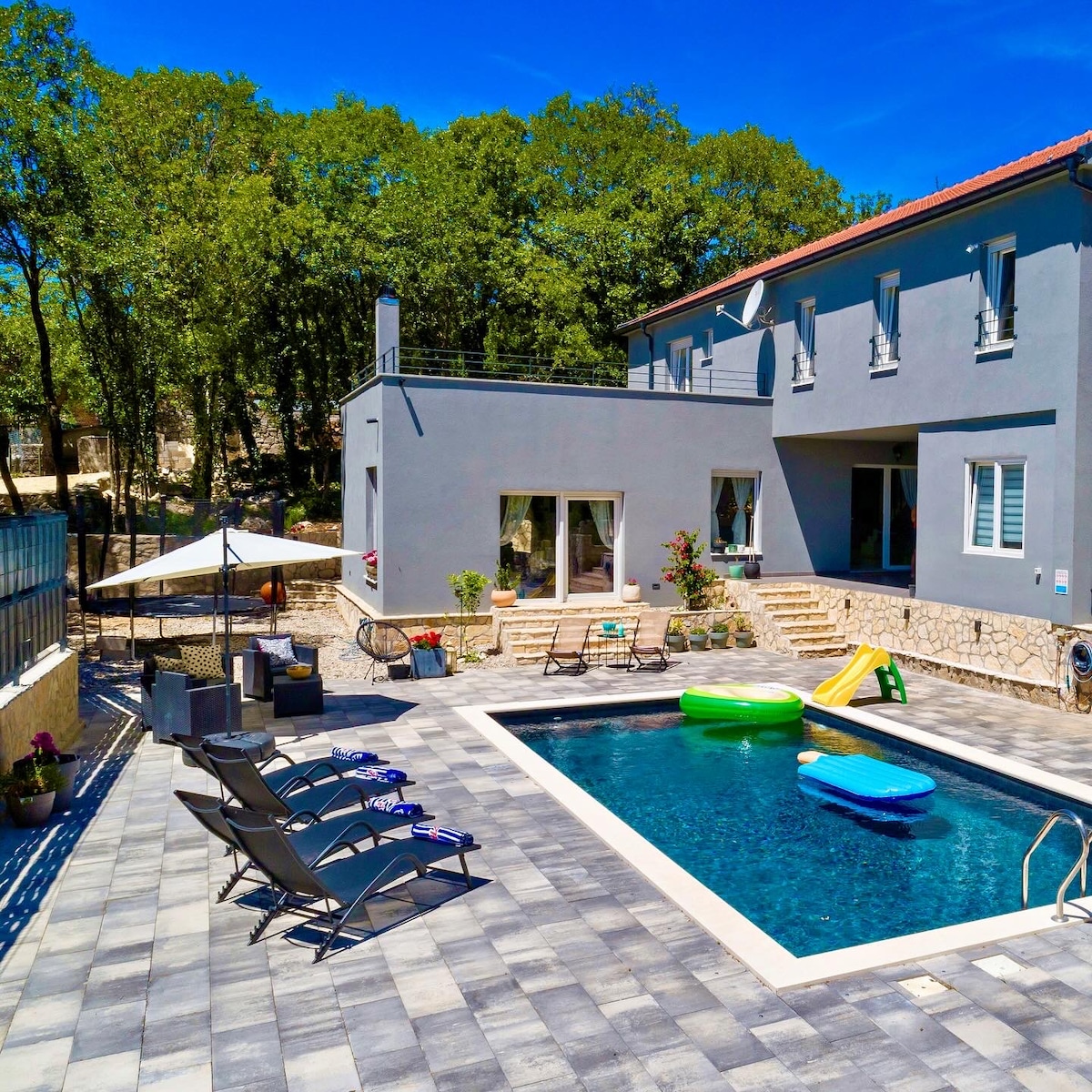 A modern villa is showcased with a spacious patio surrounding a pool. Lounge chairs are arranged neatly by the water, while colorful pool floats rest in the pool. The background features lush green trees, enhancing the serene outdoor environment.