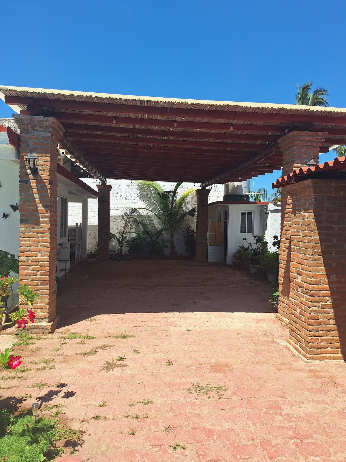 An outdoor shaded area is featured, supported by sturdy brick columns and a wooden beam roof. The floor is paved with rustic red tiles, while greenery, including a palm tree and flowering plants, adds a natural touch to the serene environment.