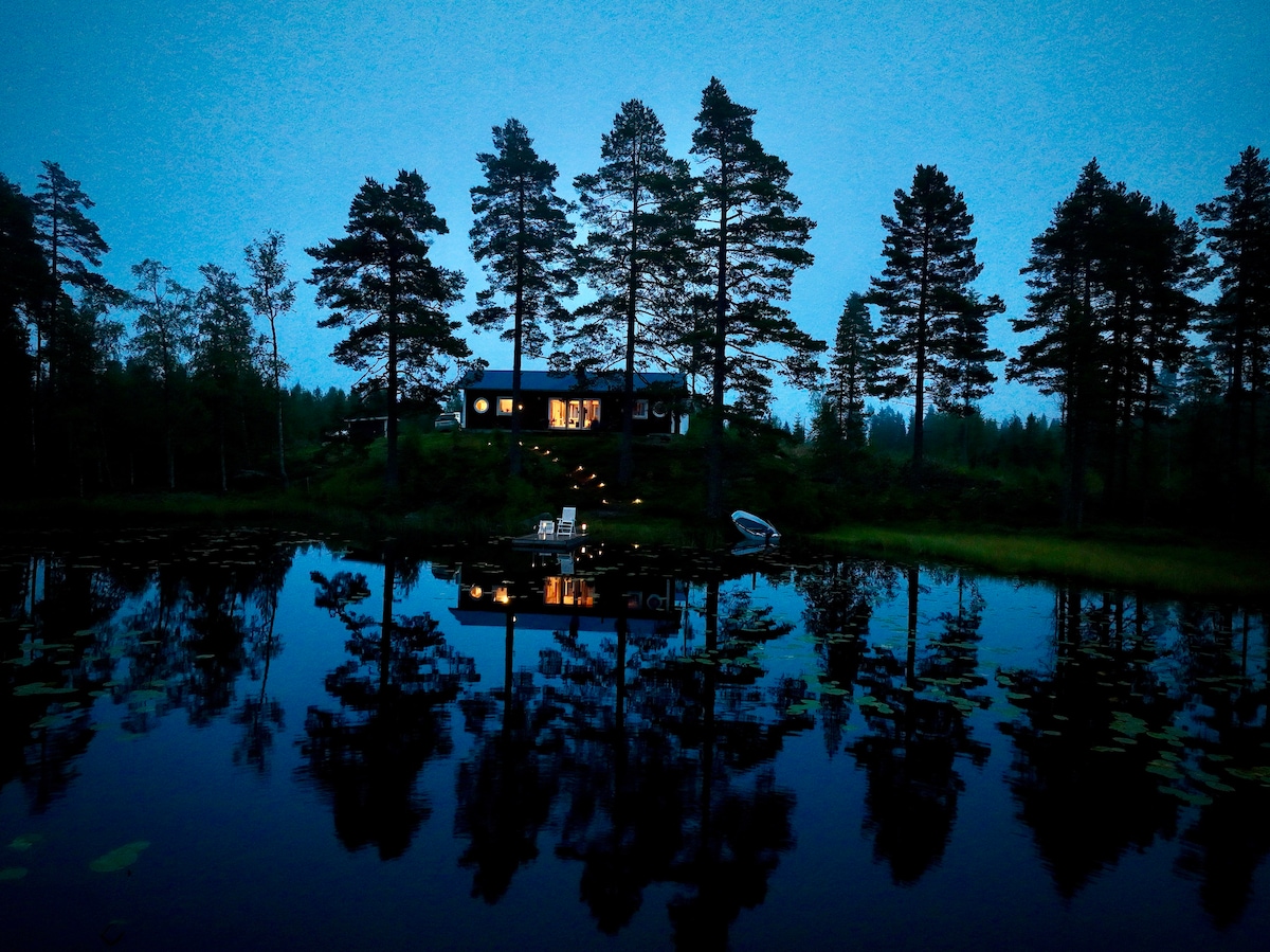 A secluded cabin is situated by the water's edge, surrounded by tall pine trees. The evening sky casts a soft blue hue over the scene, reflecting the cabin's warm lights and the silhouettes of the trees on the calm lake surface.
