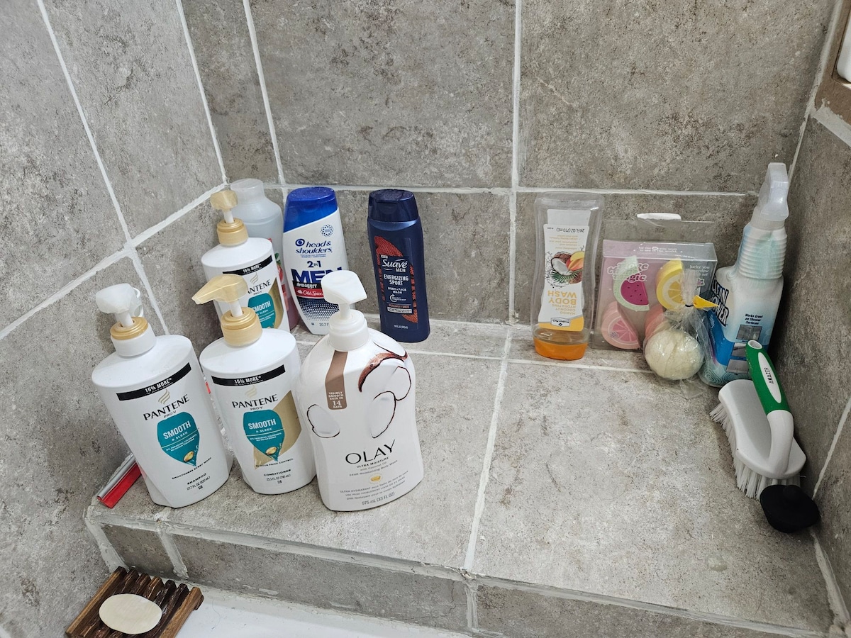 Various toiletries can be seen arranged on a tiled bathroom shelf. Several bottles of shampoo and body wash are displayed, alongside lotions and a bar of soap. A small scrub brush and a few travel-sized items are visible, providing convenience for guests.