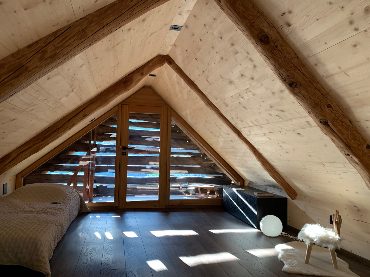 The sleeping area features sloped ceilings adorned with wooden beams, creating a spacious feel. A large window allows natural light to illuminate the space, showcasing a simple bed covered with a light blanket. A soft rug and a decorative balloon complement the serene environment.