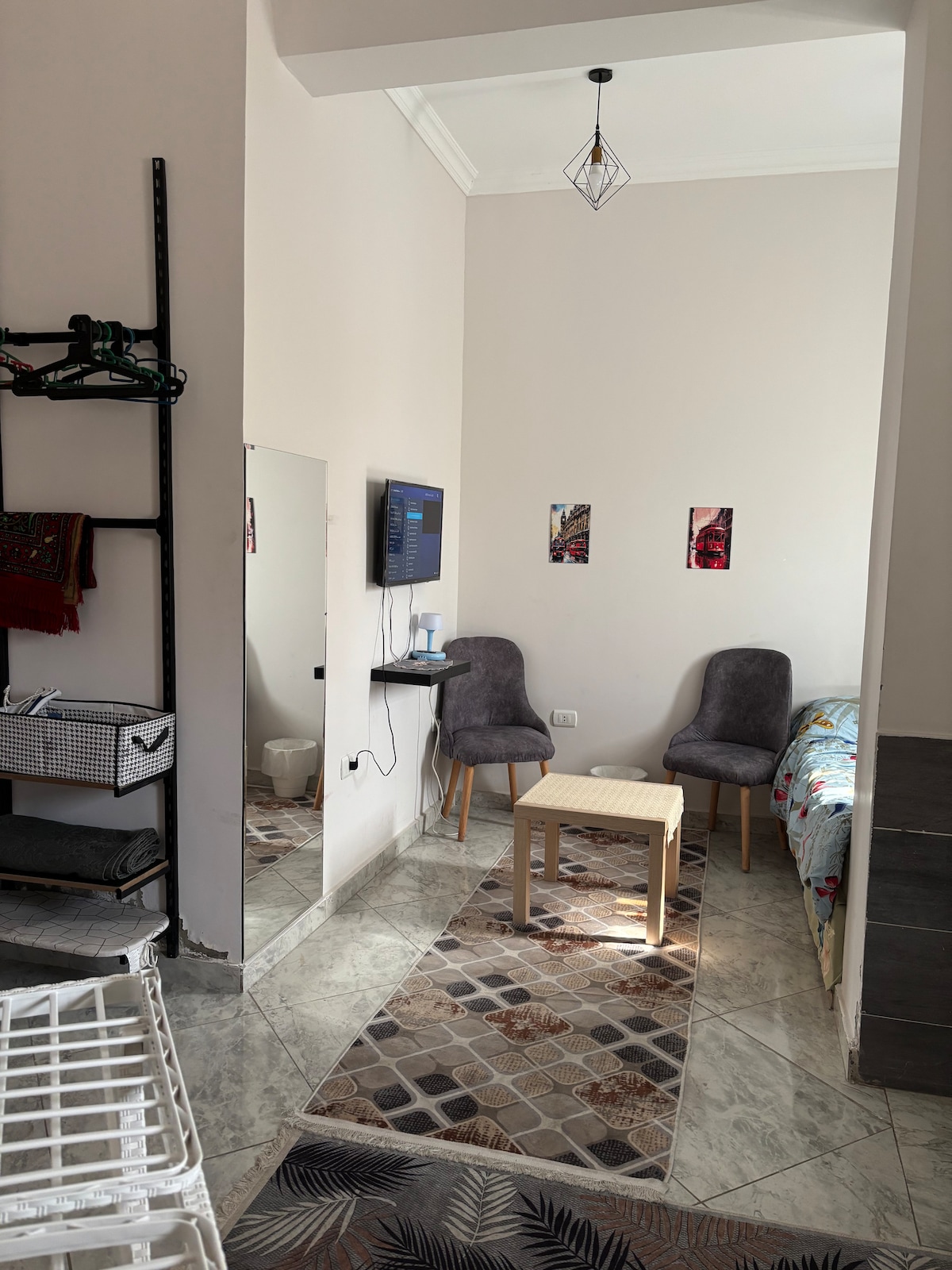 A studio apartment is displayed, featuring a small seating area with two gray chairs positioned beside a light wooden table. A mounted TV is visible on the wall opposite, and a mirrored wardrobe reflects the room's natural light. Decorative artwork adorns the walls.