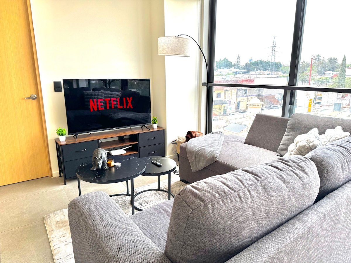 A modern living area features a large gray sectional sofa and a round black coffee table. A flat-screen TV displays the Netflix logo against a backdrop of natural light, coming through expansive windows with views of the surrounding landscape.