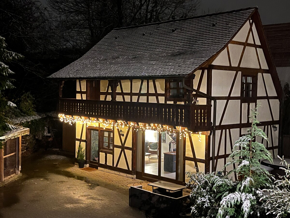 The charming timber-framed property is illuminated by soft lights, creating a welcoming presence against the snowy backdrop. A balcony is visible on the upper level, while the inviting entrance showcases a spacious area inside, viewed through opened doors.