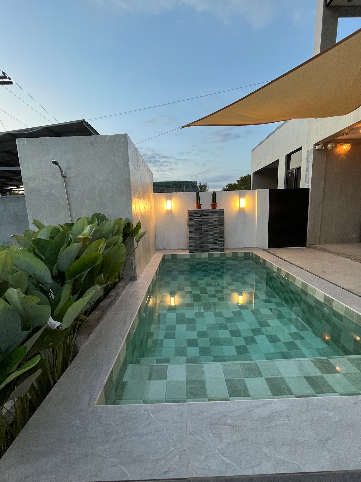 A plunge pool is surrounded by lush green foliage, providing a refreshing escape. The water features a playful pattern of light and dark tiles, reflecting the soft glow of wall-mounted lights and natural light from the sky.
