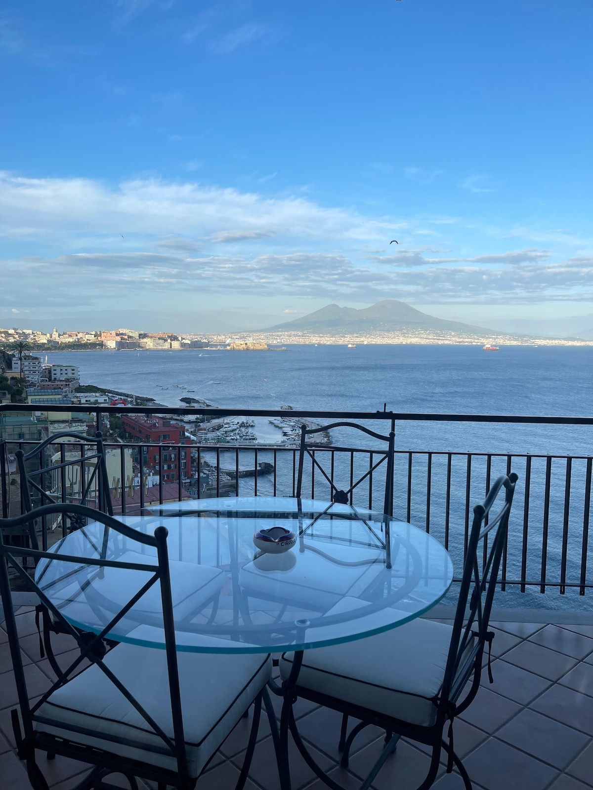 A glass table with four black metal chairs is positioned on a balcony overlooking the water. The view includes a distant volcano and the coastline, with boats visible in the harbor against a backdrop of blue skies and scattered clouds.