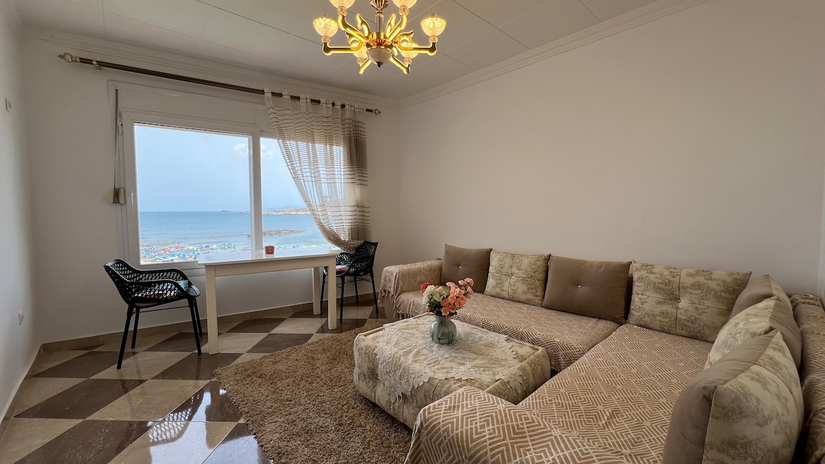 A bright living area features a spacious sectional sofa adorned with neutral-toned fabrics. A low coffee table holds a floral arrangement. Large windows offer views of the beach and distant water, while a stylish light fixture illuminates the room.