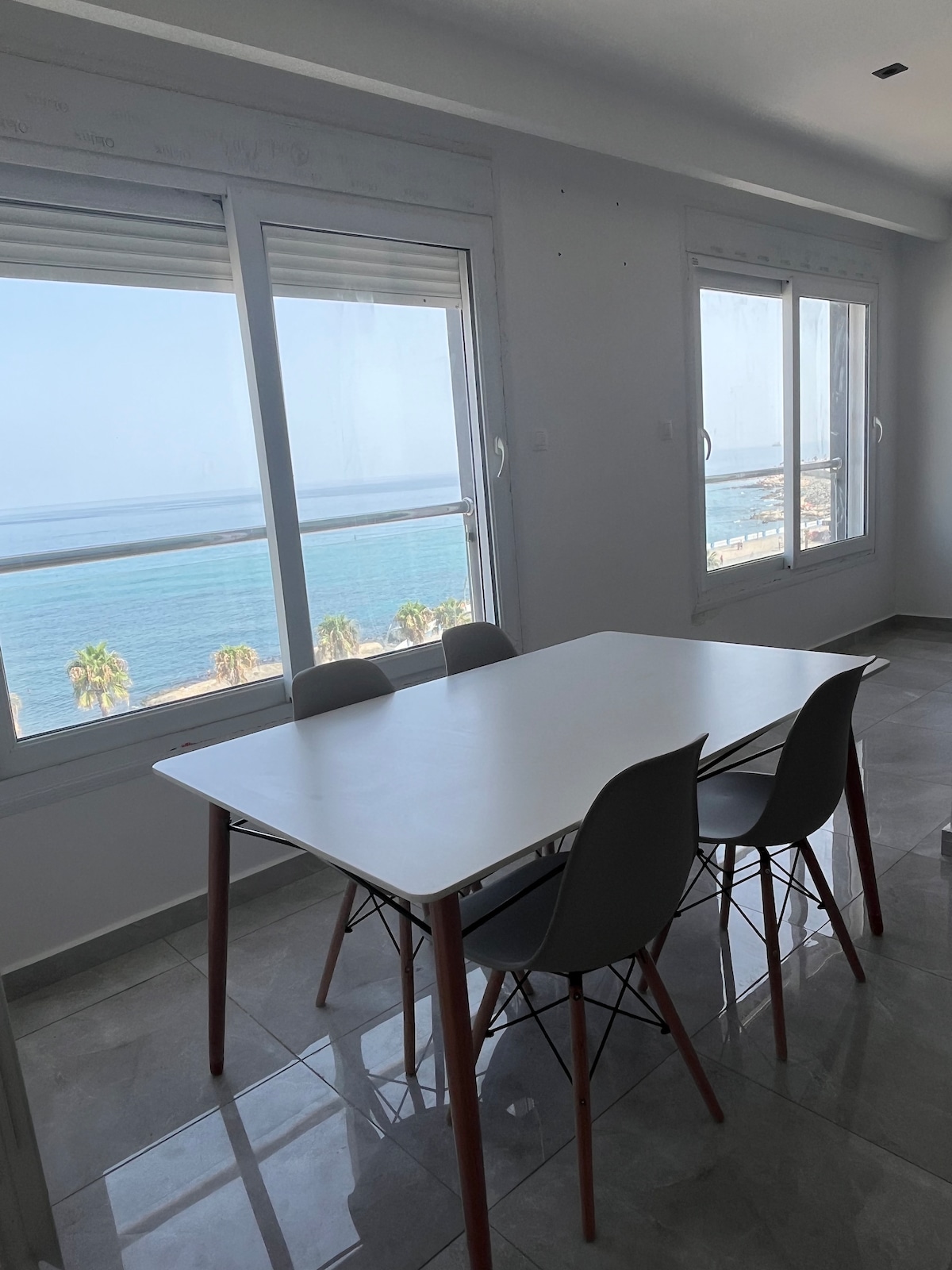 A dining area is positioned by large windows offering sea views, allowing natural light to fill the space. A white table is complemented by four grey chairs, creating a bright and open atmosphere.