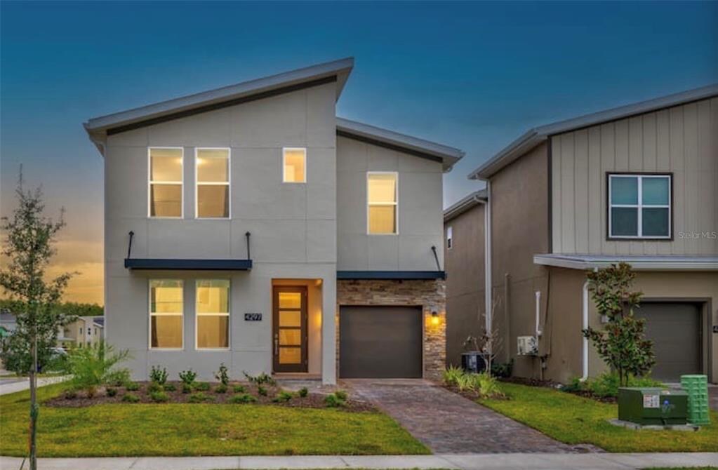 The modern two-story house features a clean exterior design with a mix of light-colored stucco and stone accents. Large windows allow natural light to fill the interior. A landscaped front yard with small greenery and a brick pathway leads to the front door.