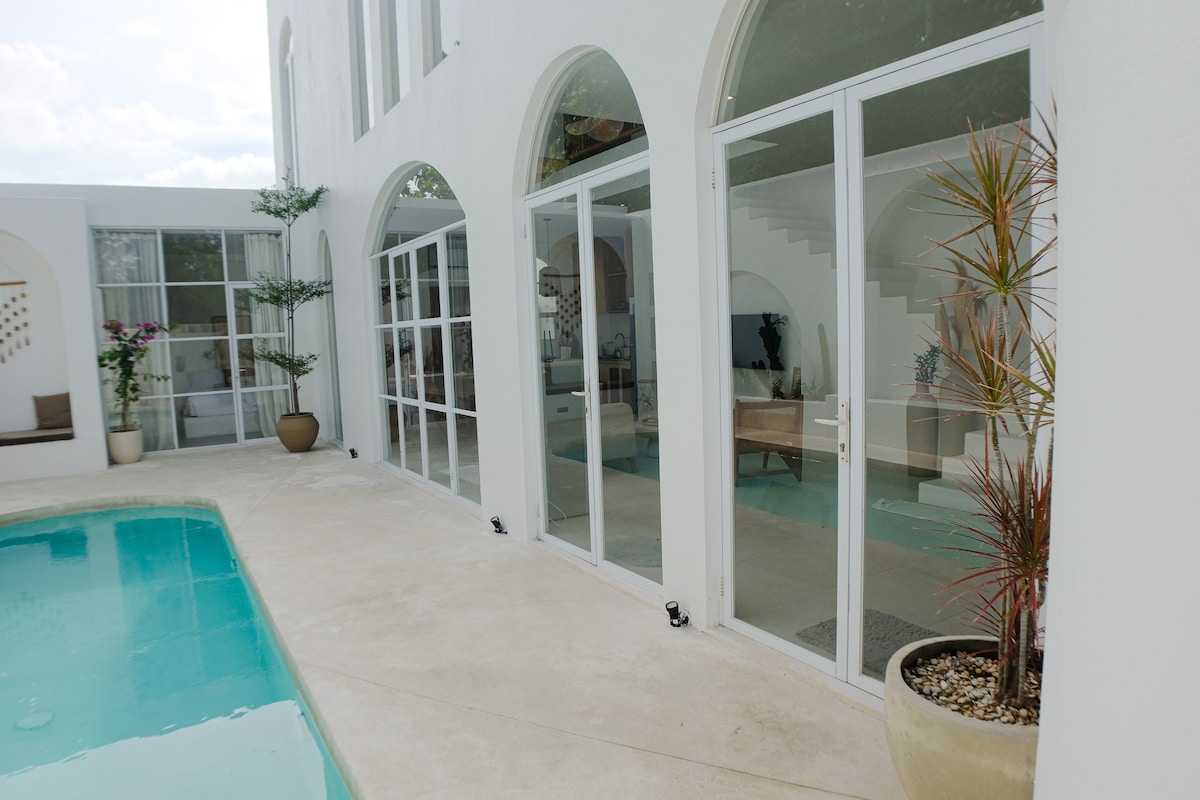A serene outdoor area features a private pool beside a spacious terrace. Large glass doors, framed by arched openings, provide a view into the living space, where light colors and minimalistic decor can be seen. Potted plants add a touch of greenery.