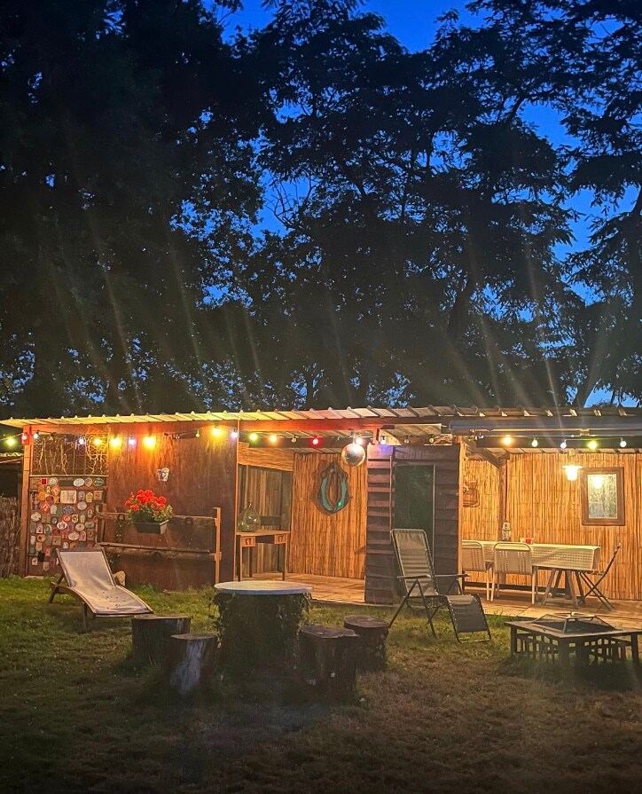 A charming outdoor space is illuminated by string lights, highlighting the wooden structure of the gîte. A seating area with lounge chairs is surrounded by a grassy garden and natural wooden stumps, offering a relaxing environment for gatherings or meals.