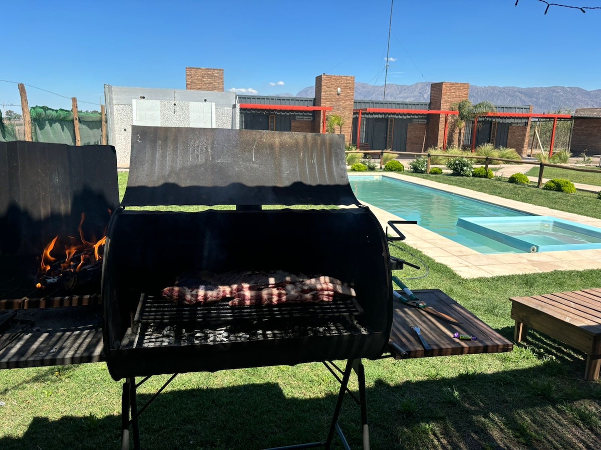 A barbecue grill is positioned in the foreground, featuring meat cooking over an open flame. In the background, a well-maintained pool is visible, surrounded by grassy areas and lounge seating. Modern accommodations are seen beyond the pool under a clear blue sky.