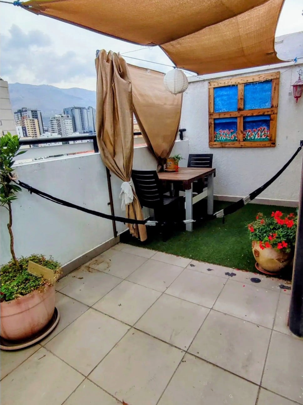 A private terrace is depicted, featuring a table and chairs accompanied by greenery. A decorative framed painting and a hanging lantern add character to the space, which is shielded from sunlight by a canopy. City views are visible in the background.