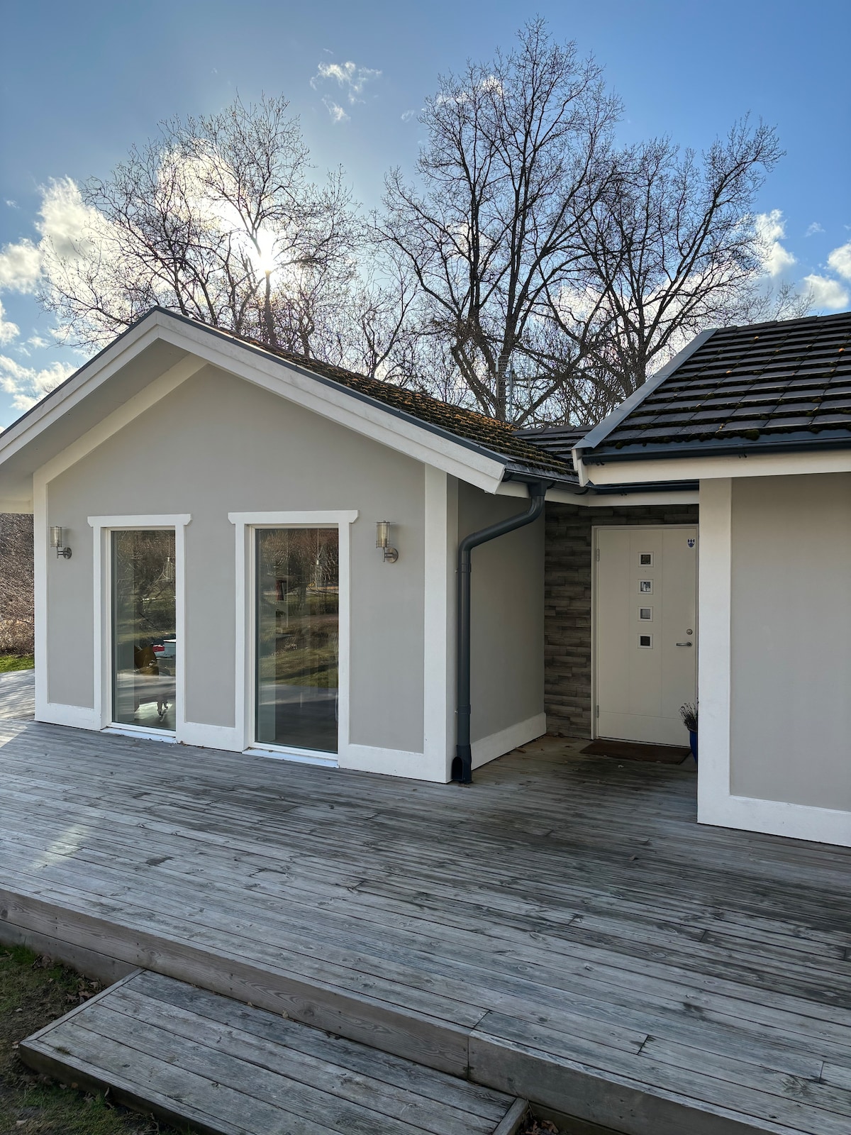 A modern summerhouse exterior is featured, characterized by a light grey facade and white trim. Large glass doors provide access to a spacious wooden terrace. The structure is framed by trees and is complemented by a bright blue sky with scattered clouds.