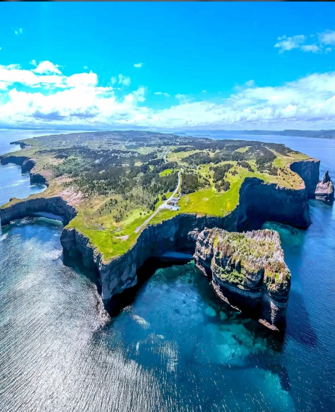 An aerial view captures the striking coastline of an island, showcasing rugged cliffs that rise above clear blue waters. Lush green grass covers the land, with winding paths visible along the shore. The vast ocean extends beyond, meeting the horizon under a bright sky.
