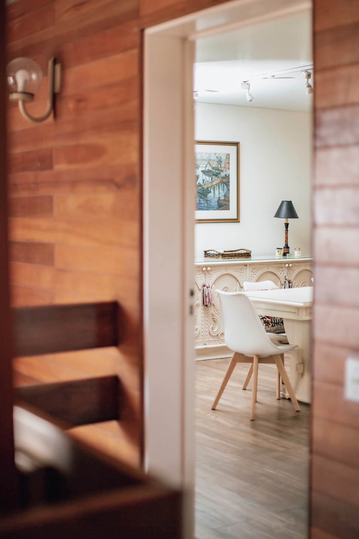 A wood-paneled entrance leads to a bright study area featuring a desk and a white chair. A desk lamp and framed artwork add a touch of personality, while natural light enhances the warmth of the wooden surroundings.