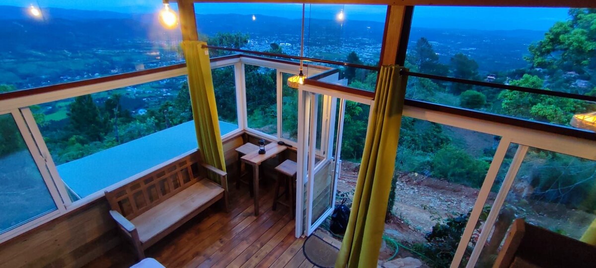 A bright interior features large windows that offer panoramic views of the surrounding landscape. Simple wooden furniture includes a small table and a couple of chairs, while vibrant green curtains frame the windows, enhancing the natural light that fills the space.