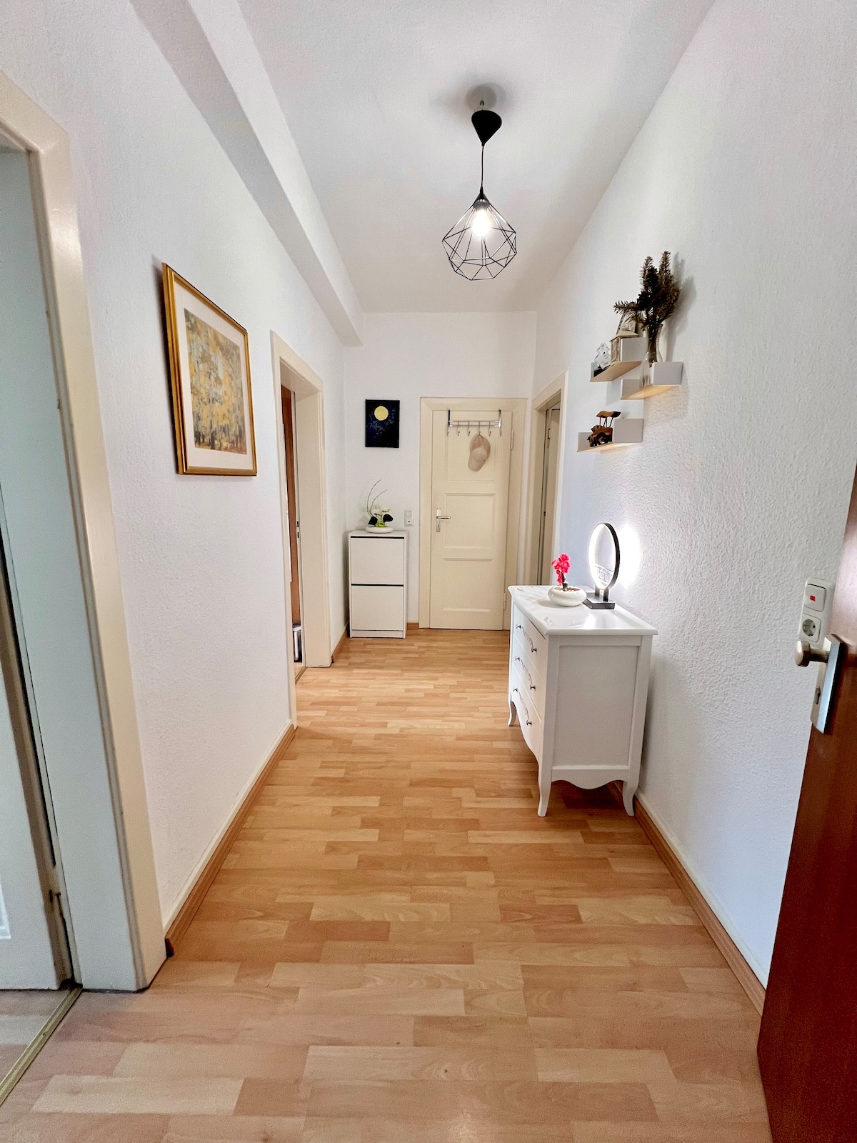 A well-lit hallway is presented, featuring light wood flooring and white walls. A decorative mirror and a small vase with flowers can be seen on a white console table. Doors to various rooms are visible along the hallway, contributing to a spacious feel.