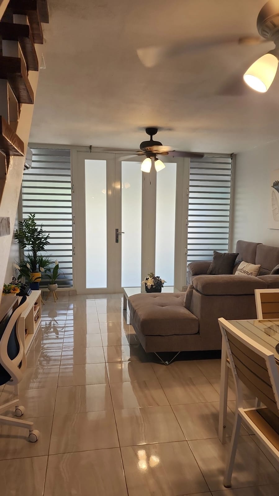 The living area is furnished with a spacious sectional sofa and a coffee table, illuminated by natural light from glass doors. A dining table and desk are positioned nearby, with greenery adding a refreshing touch to the space. The smooth tiled floor reflects the light, contributing to an open feel.