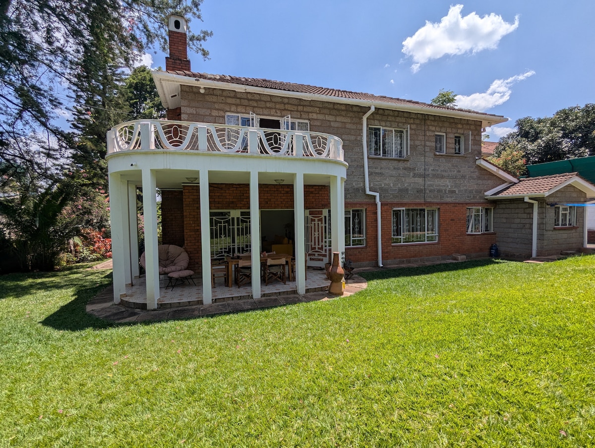 A spacious exterior view of the villa is presented, featuring a covered terrace supported by white columns. The lush green lawn surrounds the structured exterior, which includes multiple windows and a cozy seating area. Clear blue skies and scattered clouds provide a serene backdrop.
