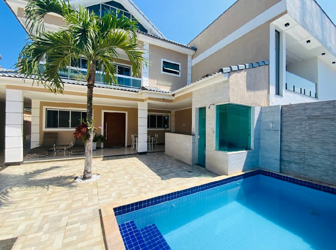 The outdoor area features a private, unheated swimming pool surrounded by a tiled patio. A palm tree provides shade near the pool area. The two-story house showcases large windows and doors, enhancing the bright, sunny atmosphere.