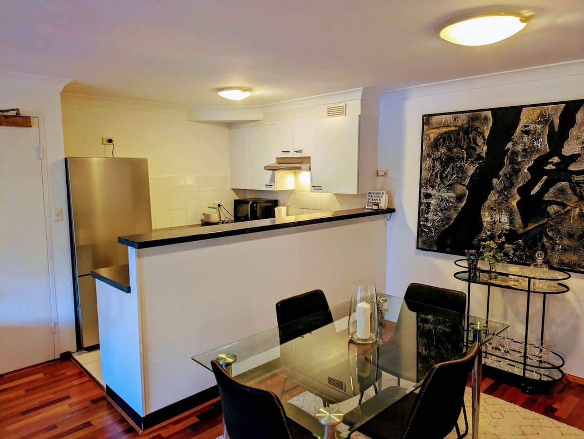 A modern kitchen is visible, featuring stainless steel appliances and a dark countertop. A dining area with a glass table and black chairs is positioned nearby. An artwork hangs on the wall, adding character to the space.