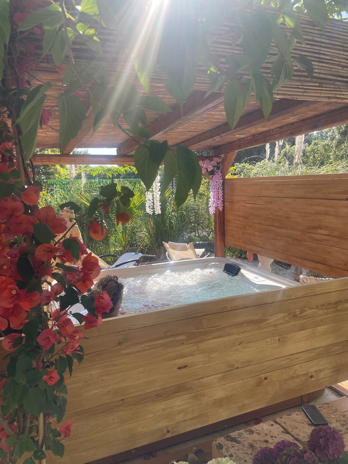 A hot tub is situated under a wooden pergola, surrounded by vibrant flowers and lush greenery. Sunlight filters through the leaves, adding warmth to the serene outdoor setting. Comfortable lounge chairs are placed nearby, enhancing the relaxing ambiance.