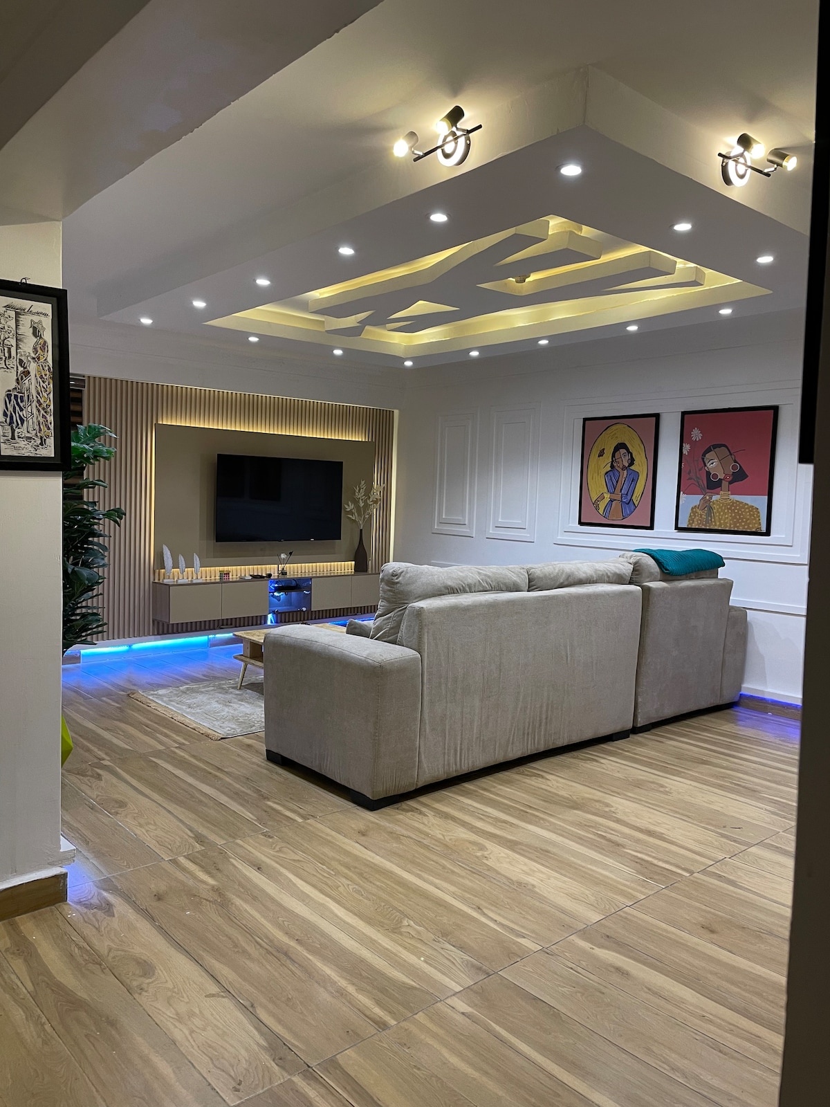 A modern living area features a comfortable sofa, decorative wall art, and a wall-mounted television. Ambient lighting accentuates the ceiling design and shelves. Wooden flooring complements the overall aesthetic, while blue LED lighting adds a contemporary touch.