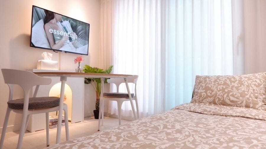A cozy room is equipped with a smart TV mounted on the wall, displaying content. A table is positioned next to the bed, accompanied by two chairs. Light filters through sheer curtains, creating a bright atmosphere in the space.