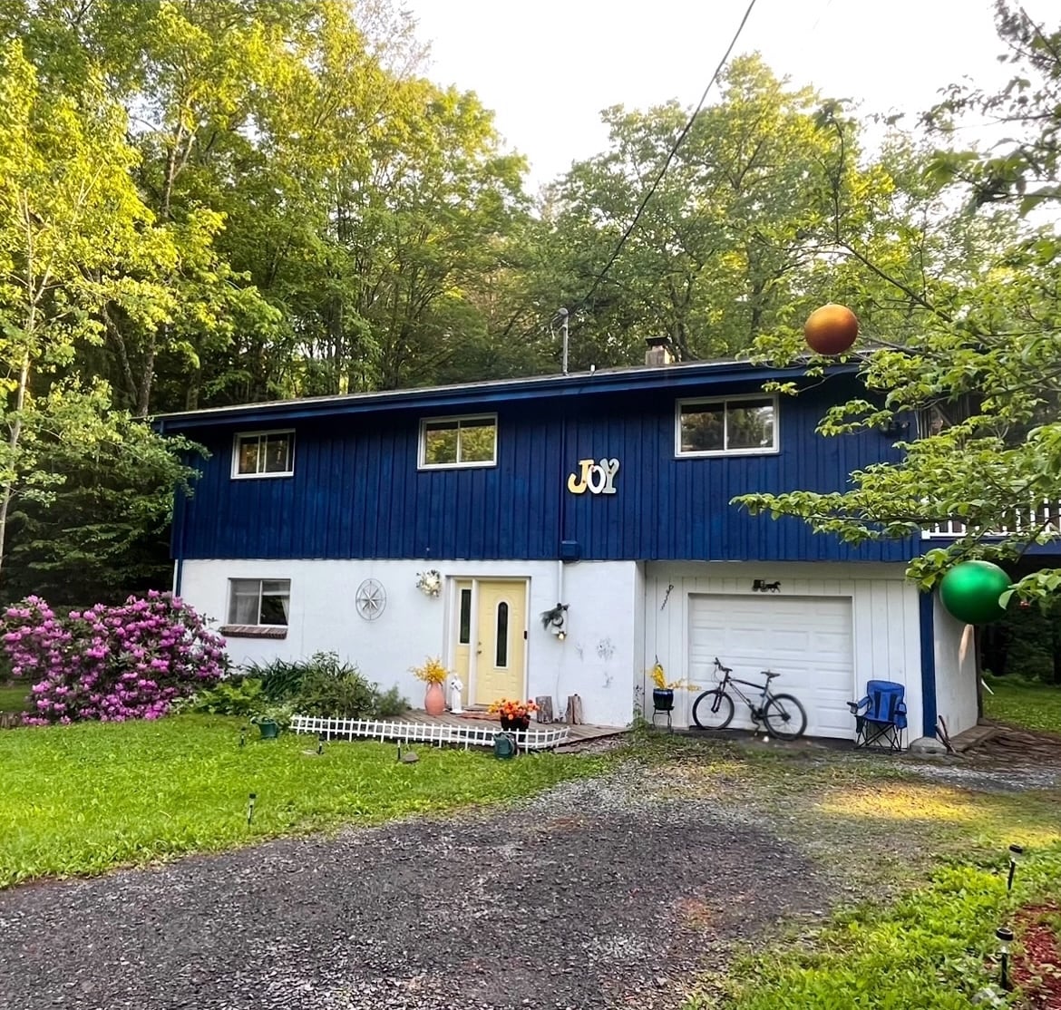 A ranch-style house is set among lush greenery, characterized by a blue exterior and white trim. A gravel driveway leads to the entrance, with a blooming azalea bush adding color. A bike is parked near a blue chair, suggesting outdoor leisure opportunities.
