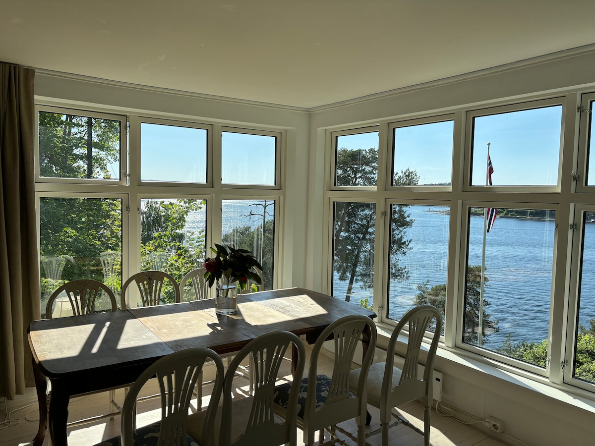 A spacious dining area is bathed in natural light from large windows that overlook the water. A wooden table is set with chairs around it, and a potted plant adds a touch of greenery. The view includes trees and the fjord beyond.