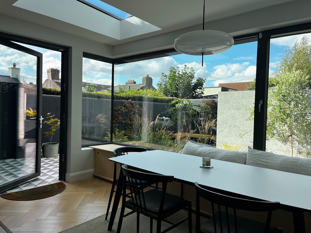 A bright dining area features a long white table surrounded by black chairs. Large glass windows offer views of a landscaped garden. A skylight allows natural light to fill the space, and a dark-framed door opens to the outdoor area, adding to the openness.