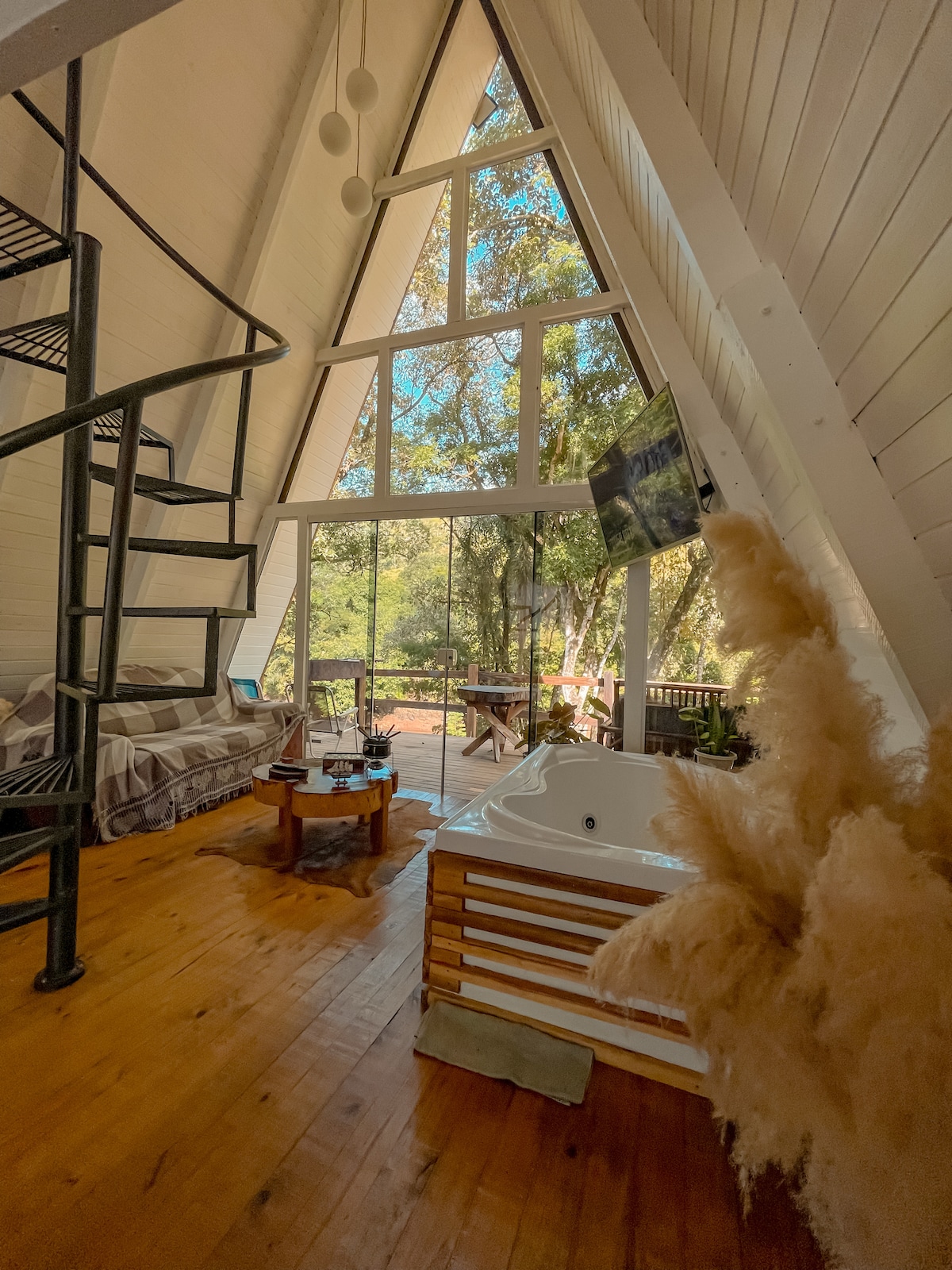 An A-frame interior is visible, featuring tall windows that invite natural light and display views of the surrounding greenery. A cozy seating area is arranged near a wooden table, while a hot tub is positioned nearby. A spiral staircase leads to the upper level.