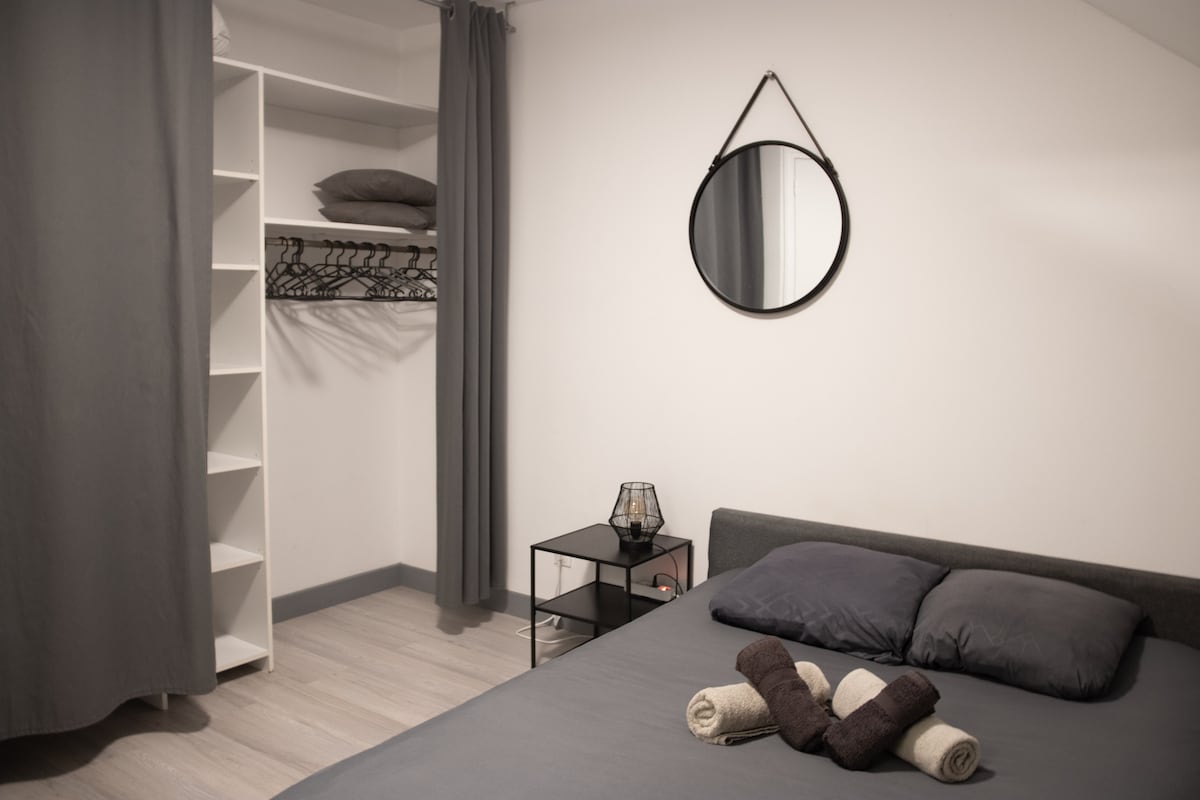 A well-lit bedroom features a spacious bed with dark linens and neatly rolled towels arranged on the bed. A round mirror is positioned above a small side table, and a closet with shelves is visible in the background.