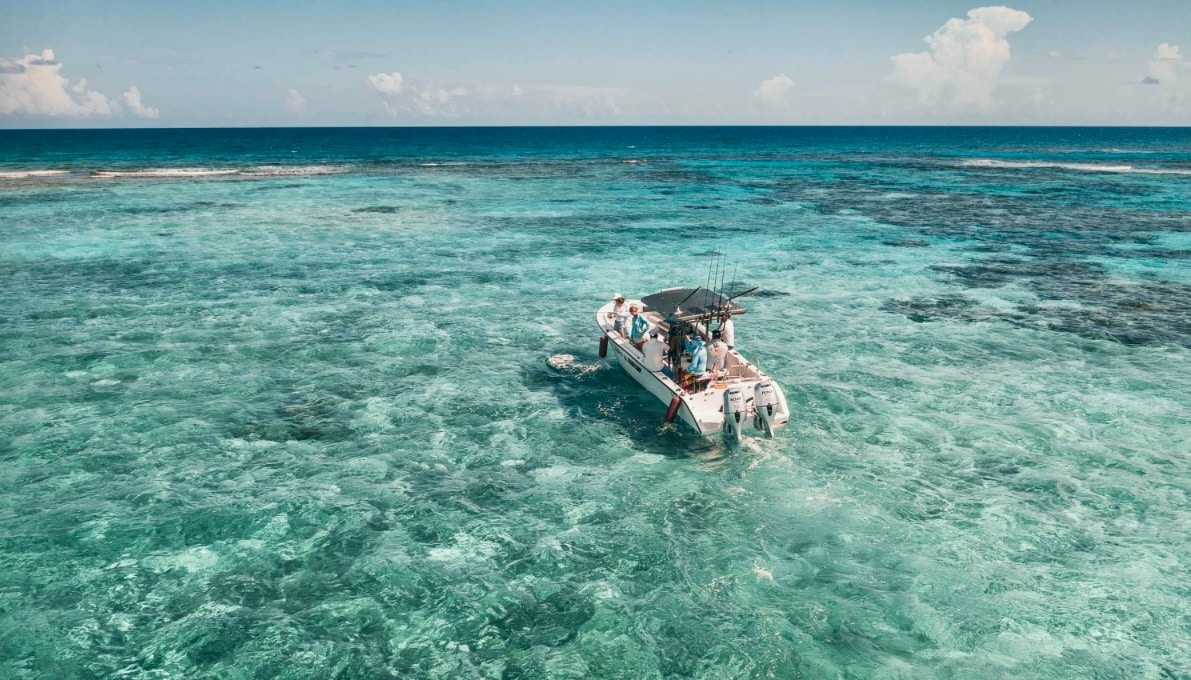 A boat is anchored in clear turquoise waters, surrounded by vibrant coral reefs. The sun glimmers on the water’s surface, creating a serene atmosphere. Cloud formations are visible in the blue sky above, adding to the peaceful coastal scene.