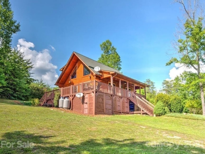The inviting log cabin is set on a spacious green lawn, surrounded by trees. A large deck extends across the front, offering ample outdoor space. Windows frame the structure, providing natural light, while a staircase leads to the entrance.